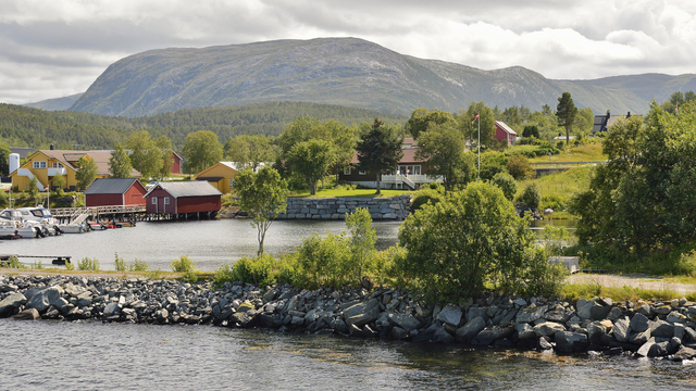 A photo of some buildings on the shore of a small town. There are low green mountains in the distance. The sky is filled with clouds.