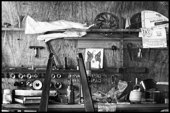 A cluttered workbench featuring various tools and hardware, including wrenches, containers, and a jar. The back wall displays a drawing of a dog, alongside keys and papers. The image is in black and white.