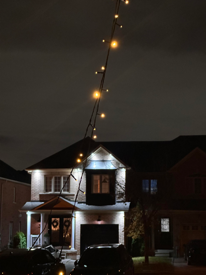 String of yellow lights hanging against a house with white outside lighting, under a snow-filled night sky.