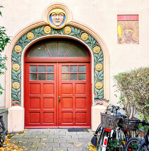A stunning Art Nouveau (Jugendstil) entrance in Munich, featuring a wooden double door with a reddish-brown finish. The door is set beneath a rounded archway decorated with a prominent frieze of stylized green foliage and golden-yellow sunflowers. Above the arch, a striking sculpted mask with a yellow face, blue headpiece, and stern expression is centered. To the upper right, a square relief carving depicts a figure in profile, possibly a sun god, holding a yellow spear. Two bicycles with wicker baskets are parked to the right of the doorway, partially obscured by green hedges. The ground is paved with stone tiles covered in fallen autumn leaves.