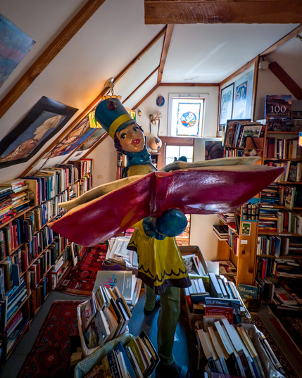 Standing on a table of books (in a room full of books), a fiberglass figure of a woman in marching band uniform that is yellow and green is holding out an open book to the viewer