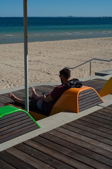Auto-generated description: A person is relaxing on colorful lounge chairs under a shade by the beach, with a view of the sandy shore and the ocean.