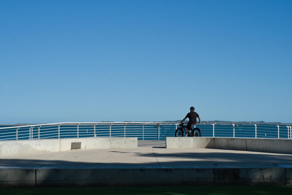 Auto-generated description: A person on a bicycle is gazing at the expansive ocean from a waterfront promenade under a clear blue sky.