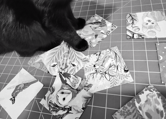 
Black and white photo of a black cat named Brannagh, sitting on top of a craft table covered with small fabric squares. The fabric squares feature various animal designs: a fox, a cat, a giraffe, a deer, an owl, and a hippo.
The table has a cutting mat-style grid pattern. 
Brannagh's position suggests she is overseeing the crafting.

