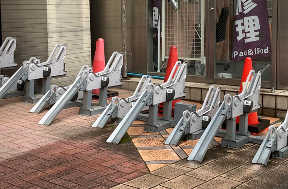 A row of metal starting blocks for bicycle parking and field positioned on a tiled surface, with orange cones placed nearby. In the background, a glass door with text.