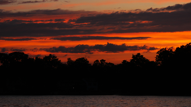 Sunset photograph of dark horizontal clouds against a red and orange background with trees and a lake below.