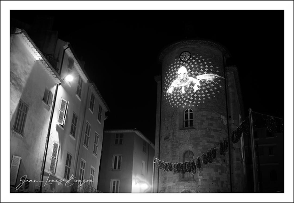 This evocative black-and-white photograph captures a nighttime scene dominated by an ancient, cylindrical stone tower. The tower commands attention with its textured, weathered facade, suggestive of centuries' worth of history. Projected onto the tower's surface is an image of a Templar Knight, rendered in a luminous, almost ethereal glow. Around the Templar Knight, a pattern of small, evenly spaced dots or stars seems to radiate, creating a celestial ambiance that contrasts strikingly with the solid stone structure.
Adjacent to the tower, the surrounding buildings are softly illuminated by street lamps, their facades marked by classic European architectural details—simple windows with shutters, sloping roofs, and stucco walls. The gentle gradient of shadows and light frames the tower as the focal point, guiding viewers to the symbolism embedded in the projection.
