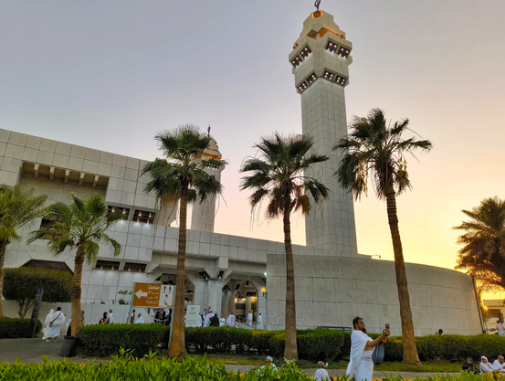 ​"A modern mosque with two tall, rectangular minarets at sunset. Palm trees stand in front of the mosque, and people in white robes are visible around the entrance and on the path. One man in the foreground is taking a photo with his phone."