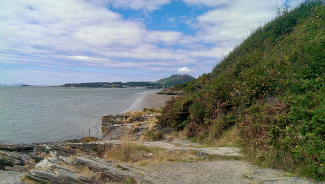 A rugged coastline stretches along a calm sea beneath a sky of scattered clouds, with gentle sunlight brightening the landscape. A narrow strip of sandy beach curves between the water and rocky outcrops, leading the eye toward distant green hills that rise softly in the background. Dense shrubs and low vegetation cover the slope on the right, contrasting with the weathered stone and dry grasses near the path in the foreground. The tide appears low, revealing wet sand and shallow water along the shore, while the horizon blends softly into the muted blues and grays of the open sea.