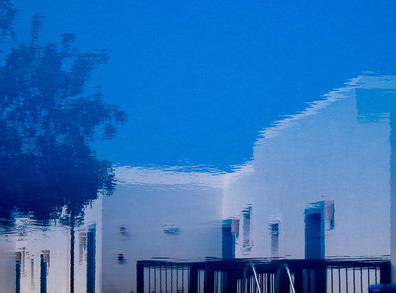 An abstract reflection of a building and trees on a water surface, creating a distorted image against a blue sky backdrop.