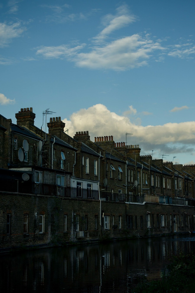 Photo of the back of a terrace of brick houses, backing onto a canal, in the shadow of a pale blue sky.