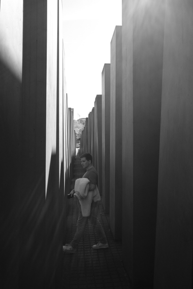 Das Schwarzweißfoto zeigt einen jungen Mann, der das Denkmal für die ermordeten Juden Europas in Berlin durchquert. Er geht auf dem gepflasterten Weg zwischen den hohen Betonstelen entlang, mit einer Jacke über dem Arm. Die Lichtstimmung wird durch das Gegenlicht und Streulicht im Objektiv bestimmt. The black-and-white photo shows a young man walking across the Memorial to the Murdered Jews of Europe in Berlin. He is walking along the paved path between the tall concrete steles, with a jacket over his arm. The lighting is determined by the backlighting and stray light in the lens.