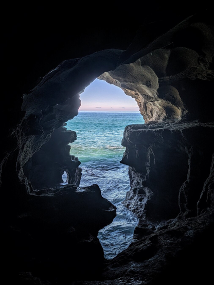 A vertical photo taken from inside a dark, rocky cave looking out through a jagged, arch-like opening. The opening frames a bright view of the turquoise and deep blue ocean meeting the sky. Sunlight illuminates the shimmering water, creating a strong contrast with the dark, textured silhouette of the cave walls on the left and right.