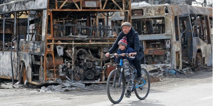 A father & son ride a bicycle past a destroyed bus.