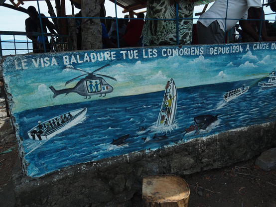 Fresque à Anjouan dénonçant le visa Balladur qui augmente les trajets en kwassa kwassa : on y voit un hélicoptère de la police française, un bateau en train de chavirer et les personnes à bord en train de se noyer ou être dévorer par un requin
