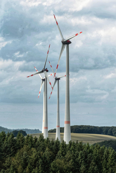 Auf dem Bild sind vier Windkraftanlagen in einer hügeligen Landschaft zu sehen. Die großen, weißen Türme mit ihren Rotorblättern, die rote Markierungen an den Spitzen aufweisen, ragen prominent vor einem bewaldeten Gebiet und einem bewölkten Himmel empor.