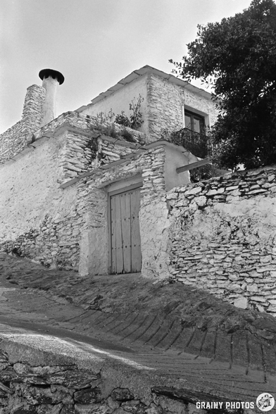 Black and white photo of an old stone house on a sloping street in Capileira. The house has a wooden door, chimney, small balcony, and rough stone walls; a tree’s branches extend over the building.