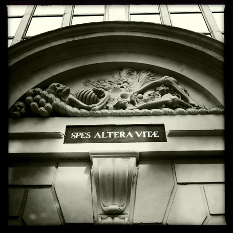 Moody monoschrome photo of an old ornament on an Amsterdam building, featuring a sculpture of a lying skeleton. Underneath is the text "Spes Altera Vitae".