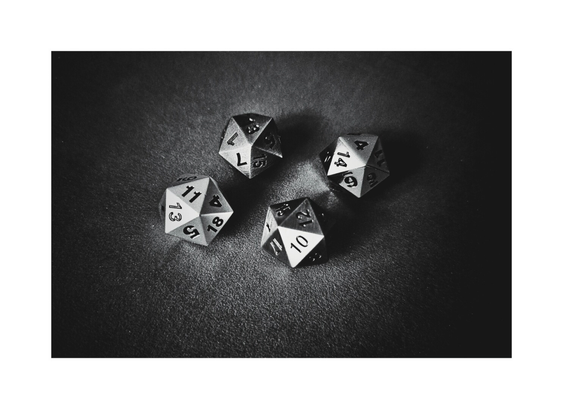 A high-angle, close-up, black and white photograph of four metallic twenty-sided dice (d20) resting on a dark, velvety surface. The dice are casually grouped, with various numbers visible on their top faces. The lighting creates strong highlights and shadows, emphasizing the sharp edges and numbering of the dice.