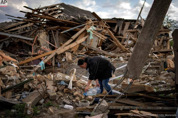 A woman watering a plant in a container in front of a destroyed house.