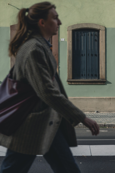 A blurred person walks past a green building with closed blue shutters and brown wooden trim, casting soft afternoon shadows on the textured wall.