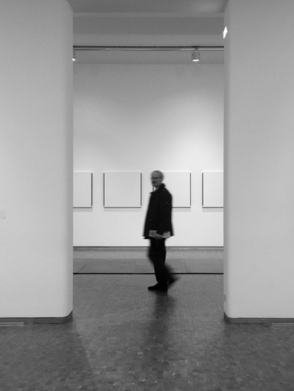 das bild zeigt eine schwarz-weiß-aufnahme eines mannes in einem museum. der mann geht von rechts nach links und ist dabei mittig im bild. er hat der kopf gedreht und blickt in die kamera. der mann wird links und rechts von zwei hohen wänden gesäumt. hinter ihm an der wand hängen vier quadratische gemälde, die teilweise dunkle ränder haben, aber sonst weiß wirken. der mann ist unscharf, da er in bewegung ist. der boden besteht aus holzparkett. die räumlichkeit wirkt minimalistisch und modern.