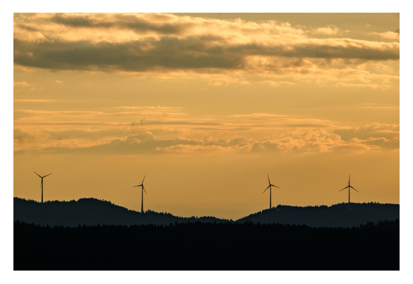 Foto im Querformat. Goldene Stunde. Unten sind die Silhouetten von bewaldeten Hügelketten. Am Horizont stehen vier Windkraftanlagen in einigem Abstand zueinander. Der Himmel ist golden mit einigen Wolken.