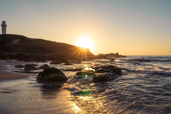 Sunrise view of Wollongong City Beach with gentle waves, golden morning light, and the Wollongong Head Lighthouse in the far distance.