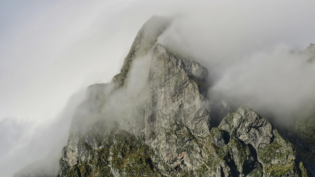 A colour photo of a mountain top surrounded by dense cloud. The peak has two distinct tops and there is cloud between them, giving the impression of being caught by them in a pinch. The rock is mostly grey but some green vegetation can be seen on horizontal ledges.
