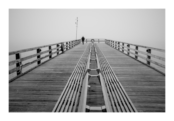 Ein SchwarzWeiß - Bild aud dem ein Mann auf einer Seebrücke geht. Das Foto ist stark symmetrisch aufgebaut, alle Linien streben zur Mitte hin