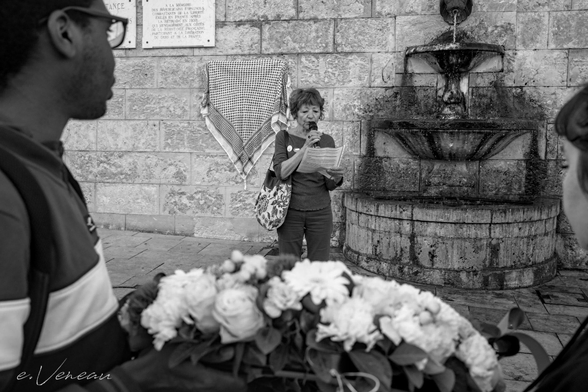 Two young people with their backs turned carry a wreath of white flowers in memory of Palestinian children, while a middle-aged woman reads a speech in front of a plaque hidden by a keffiyeh.