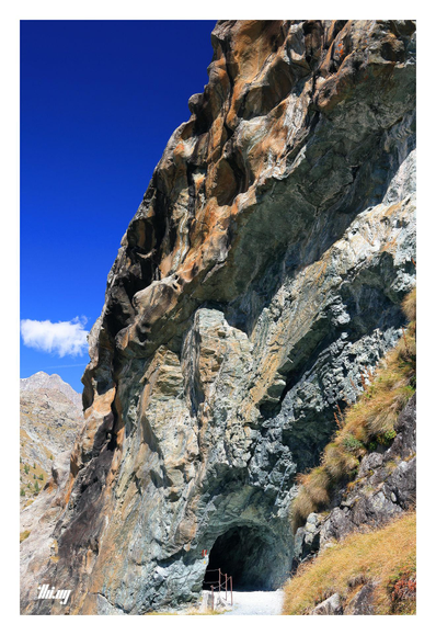 View of a small tunnel entrance (2-3 meters wide) into a glacially eroded colorful rock face consisting of slightly curved strata of different minerals and colors ranging from blueish-green grays to rusty orange. The higher up sections show signs of glacial plucking and trigger the imagination how that place must have looked filled with ice... A tall mountain peak in the far back. Clear, deep blue sky.