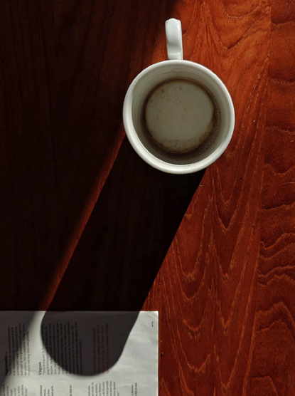 A photo looking down on a white mug with coffee residue, sitting on a wooden table and casting a long shadow that falls across a newspaper page.