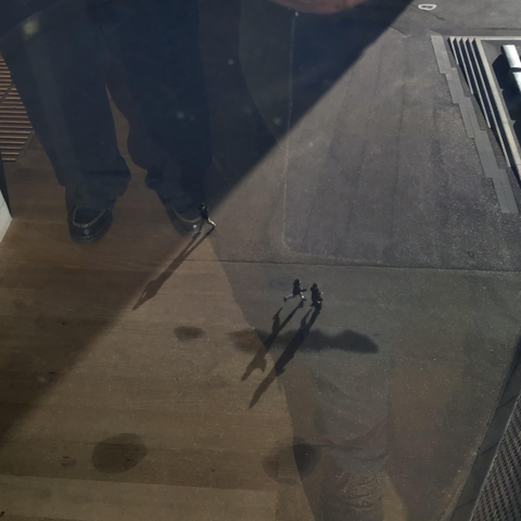 A photo taken from the top floor of Tate Modern looks to the long shadowed small people passing below. Top left of image are the large legs and shoes of my son reflected in the window stood next to me.