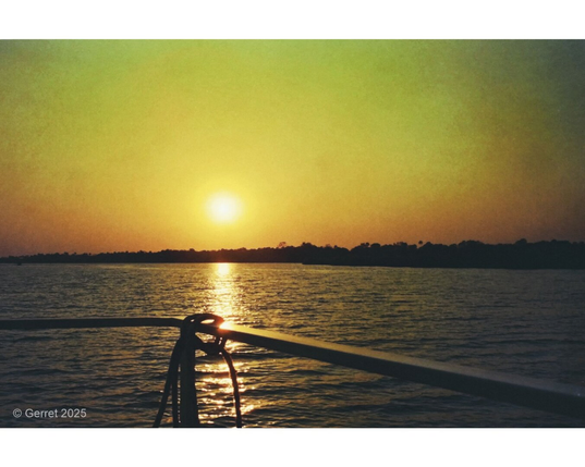 Sunset over calm waters, casting a golden glow. Silhouette of a distant shoreline and boat railing in the foreground create a serene mood.