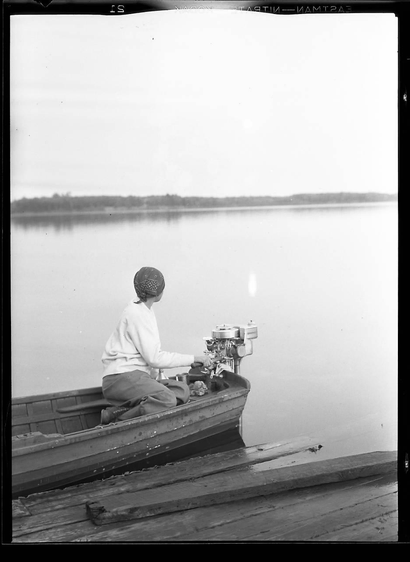 A woman in a small boat in the evening (?), watching across a calm lake. Her hand is on the handle of the outboard motor. She’s wearing a headscarf and is dressed in plain clothes. The Guardian byline: ’ Florence Carr, Fujita’s wife, on Manitowish Lake in Vilas County, Wisconsin in 1930. Fujita and Carr married in 1940 ’