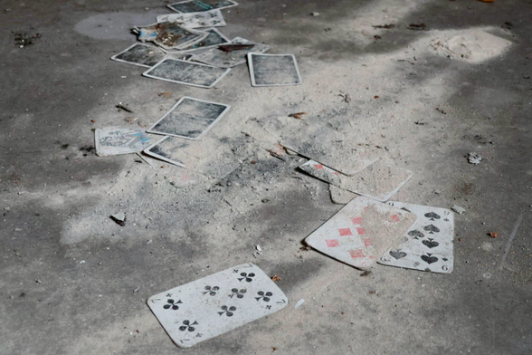 Spielkarten eines Kartenspiels liegen auf einem grauen Betonboden. Die Karten sind mit Staub überzogen und liegen entweder mit dem Deckblatt nach oben oder unten. Die Lage der Karten wirkt als wenn sie vom Tisch dorthin geweht wurden. 