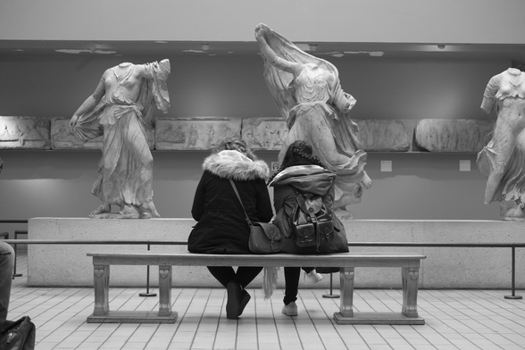 Photograph of Acropolis sculptures in the British Museum. In front of the pieces two young people are sitting on a bench with their backs to the camera. They are wearing winter coats and are carrying handbags and a leather rucksack. One has her head down as if looking at a phone. The other is looking at the sculptures. The shape of the hood on her coat echoes the shape of stone drapery, the bottom of the figure's dress on its upturned leg, on the sculpture in front of her. The three sculptures are missing heads and one arm and hand each. A frieze of sculptural reliefs runs on the wall behind them. British Museum, London, UK, 10 November 2016. Photo Cindy Kohtala  