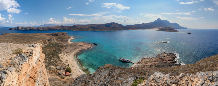Panoramic view of a coastal landscape featuring a crescent-shaped beach with clear turquoise water, a ship docked on a pier, and rugged cliffs surrounding the bay.