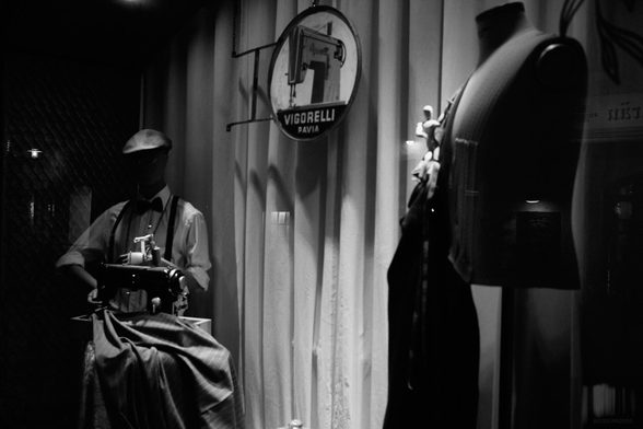 Black-and-white photograph of an illuminated shop window with mannequins, one of which is standing in front of an old sewing machine.