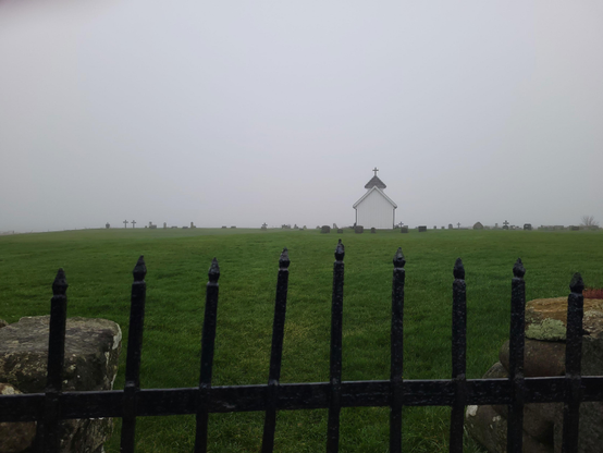 A little, white Chapel standing on a green meadow with some old gravestones surrounding it. In the foreground an iron gate sits between two stone fences. It's a foggy day and the background is all white, hiding the sea that one could only hear this time.