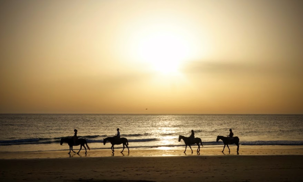 Vier Reiter am späten Nachmittag am Strand. Der Himmel ist trüb vom Meeresdunst und ggf. auch Wüstensand so dass die Sonne diffus über dem Horizont hängt. Himmel und Wasser sind in warme Farben von cremeweiß über blaßgelb bis orangebraun getaucht. Die Pferde und Personen sind als langsame Schatten auf dem nassen Sand an der Wasserlinie unterwegs. 