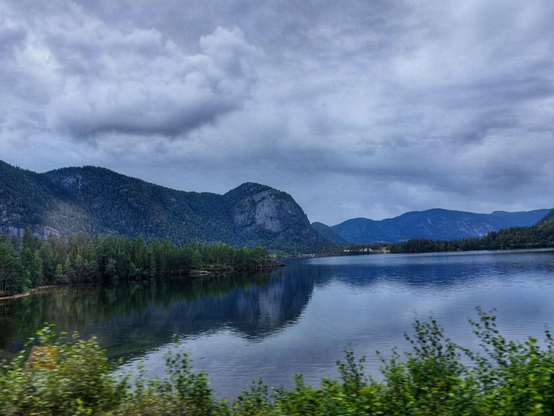 A norwegian landscape with a placid lake in the lower part of the image and mountains at the center with a cloudy sky, all reflected in the water and a green shoreline also visible
