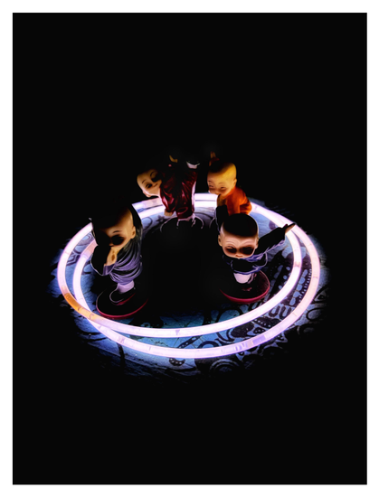 Photo of four miniature ceramic figurines of bald, baby-faced monks in different colored tunics and striking different martial arts poses. They’re grouped together inside two overlapping circles created by concert glow sticks configured into rings, on a tabletop covered in blue batik fabric. The only light comes from the glowing rings, which create striking contrasts of light and shadow on the figurines while leaving the rest of the image in darkness.