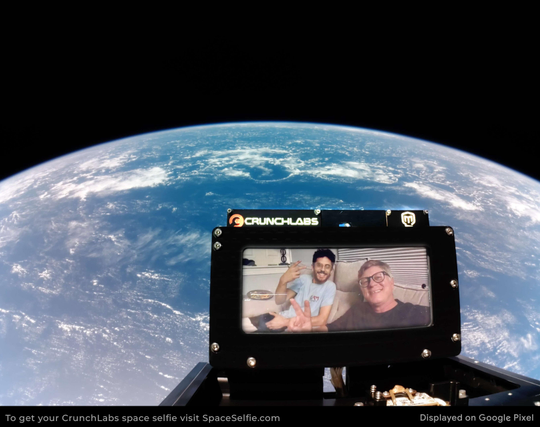 A view from space featuring Earth in the background. In the foreground, a screen displays two people posing for a selfie. The screen is branded with the "CrunchLabs" logo. The overall setting suggests a playful space-themed experience.