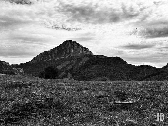 Esta es una fotografía en blanco y negro de un paisaje montañoso.
• Primer plano: Hay una zona de hierba baja y tierra rugosa que ocupa la parte inferior de la imagen. La textura del suelo es muy evidente.
• Plano medio y fondo: Domina un pico montañoso imponente con laderas escarpadas y una base cubierta de vegetación oscura (bosque o matorral). La montaña presenta zonas rocosas y acantilados.
• Cielo: El cielo está cubierto de nubes dispersas y dramáticas, lo que añade un ambiente intenso a la escena.
• Tono: El uso del blanco y negro realza el contraste y la textura, dándole a la imagen un carácter dramático y robusto.