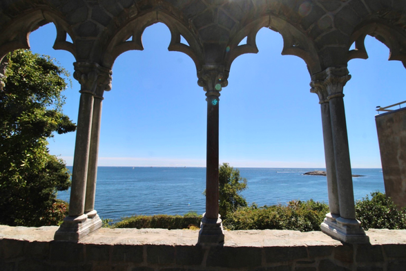 An ornate stone archway frames a serene view of the ocean, with a clear blue sky and distant boats visible. Lush greenery is situated to the left of the arch, adding to the natural beauty of the scene.