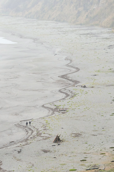 A view of a beach from high and far away. The surf is only visible in the upper left, and there are grassy cliffs along the right edge in the background. Nearby is a driftwood lean-to. In the center of the picture is a long wavy dark line at the high tide mark. There are two people waking along that line. It is raining.