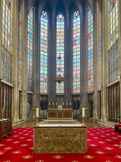 Interior of a cathedral featuring tall stained glass windows, an ornate altar in the foreground, and a richly patterned red carpet. The ambiance appears serene and majestic.
