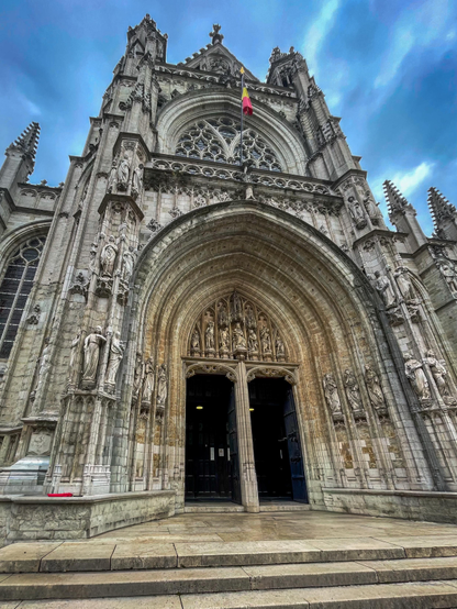A close-up view of a grand, intricately detailed Gothic church entrance, featuring a large archway with sculptures and a flag above. The facade is adorned with stone carvings, and the stairway leading up to the doors is visible.